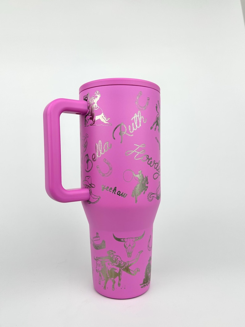 Puede incluir: Un vaso t&eacute;rmico rosa con asa, con gr&aacute;ficos plateados de tem&aacute;tica vaquera y texto, incluyendo el nombre "Bella Ruth" y la palabra "Yeehaw". El vaso est&aacute; dise&ntilde;ado para mantener las bebidas fr&iacute;as o calientes.