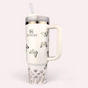 以下が含まれることがあります： ハンドル、蓋、ストローが付いた白い断熱タンブラー。タンブラーは、銀色の蝶と花の模様が特徴です。前面にはStanleyのロゴが見えます。飲み物を冷たく保つように設計されています。