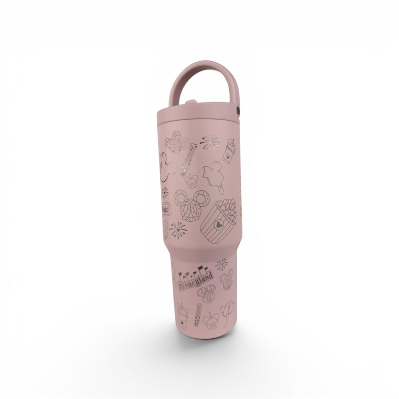 May include: A dusty rose-colored insulated tumbler with a handle on the lid, featuring a repeating pattern of etched line art icons including Mickey Mouse ears, popcorn, churros, fireworks, and the word Disneyland in a classic gothic font.