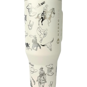 Puede incluir: Botella de agua HydroJug blanquecina con un dise&ntilde;o plateado con tem&aacute;tica de vaquero. El dise&ntilde;o incluye botas de vaquero, sombreros, caballos y la palabra "yeehaw". La botella tiene un asa y una tapa.