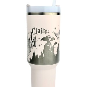 Puede incluir: Un vaso de color rosa claro con tapa y pajita blancas. El vaso presenta un gr&aacute;fico en blanco y negro de un castillo, una criatura, un b&uacute;ho y el nombre "Claire". El vaso est&aacute; dise&ntilde;ado para bebidas fr&iacute;as.