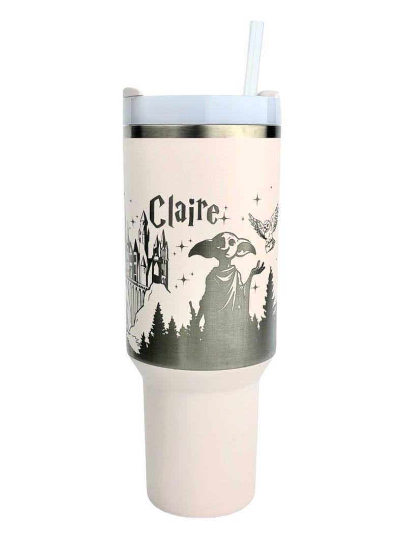 Puede incluir: Un vaso de color rosa claro con tapa y pajita blancas. El vaso presenta un gr&aacute;fico en blanco y negro de un castillo, una criatura, un b&uacute;ho y el nombre "Claire". El vaso est&aacute; dise&ntilde;ado para bebidas fr&iacute;as.
