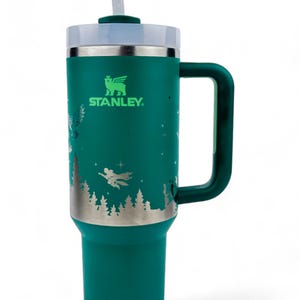 Puede incluir: Un vaso Stanley de color verde azulado con asa y pajita. El vaso presenta un dise&ntilde;o plateado de una escena de bosque con figuras voladoras. El logotipo de Stanley es visible. La tapa es blanca y la pajita es transparente.