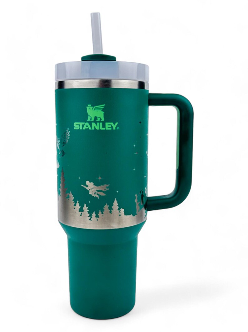 Puede incluir: Un vaso Stanley de color verde azulado con asa y pajita. El vaso presenta un dise&ntilde;o plateado de una escena de bosque con figuras voladoras. El logotipo de Stanley es visible. La tapa es blanca y la pajita es transparente.
