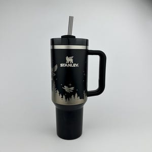 Puede incluir: Vaso Stanley negro con asa y pajita. El vaso presenta un dise&ntilde;o plateado de un b&uacute;ho, &aacute;rboles y una figura voladora. El logotipo de Stanley es visible. Dise&ntilde;ado para mantener las bebidas fr&iacute;as o calientes.