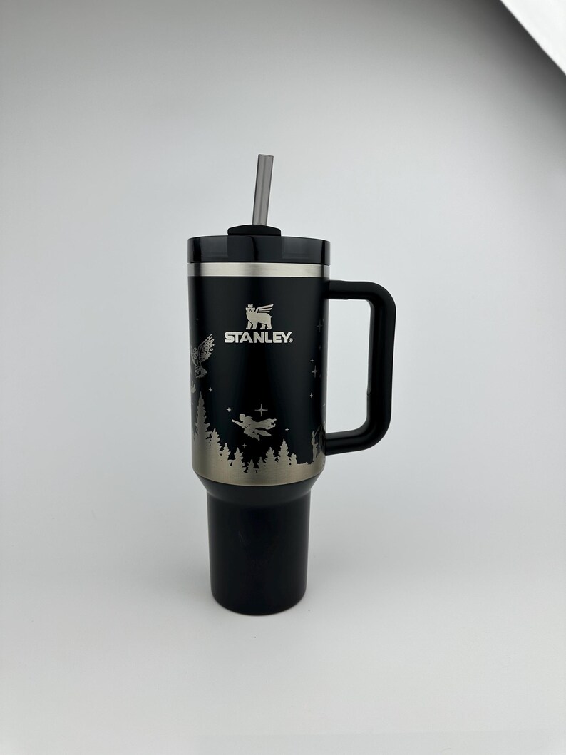 Puede incluir: Vaso Stanley negro con asa y pajita. El vaso presenta un dise&ntilde;o plateado de un b&uacute;ho, &aacute;rboles y una figura voladora. El logotipo de Stanley es visible. Dise&ntilde;ado para mantener las bebidas fr&iacute;as o calientes.