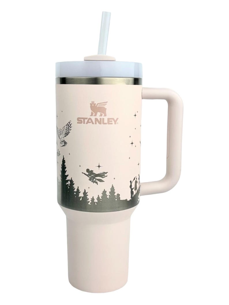 Puede incluir: Un vaso Stanley rosa claro con tapa y asa blancas. El vaso presenta un gr&aacute;fico negro de una escena de bosque con figuras voladoras y un b&uacute;ho. El logotipo de Stanley es visible. Incluye una pajita transparente.