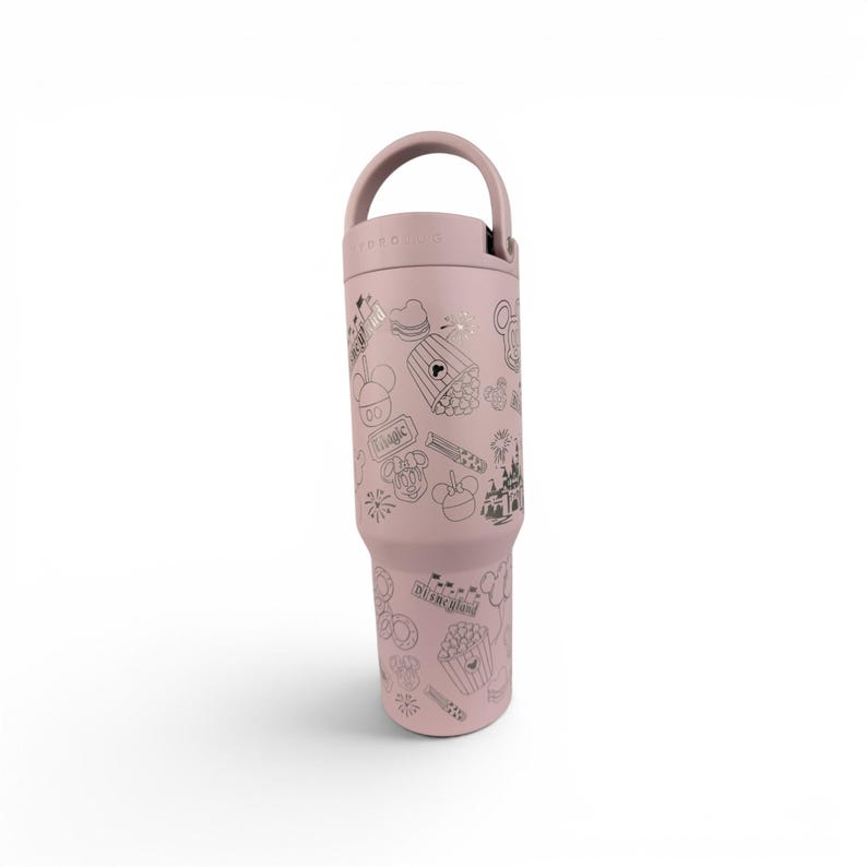 May include: A dusty rose-colored insulated tumbler with a handle on the lid. The exterior features a repeating pattern of dark gray line art illustrations including a castle, popcorn, balloons, and the text Disneyland and Magic.
