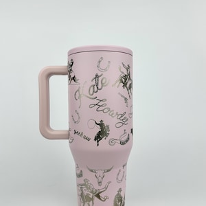 Puede incluir: Un vaso t&eacute;rmico rosa con asa, tapa y dise&ntilde;o plateado con tem&aacute;tica de vaquero. El dise&ntilde;o incluye caballos, vaqueros, herraduras y las palabras "Kate" y "Howdy".