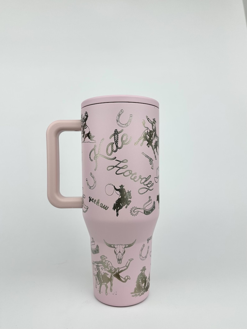 Puede incluir: Un vaso t&eacute;rmico rosa con asa, tapa y dise&ntilde;o plateado con tem&aacute;tica de vaquero. El dise&ntilde;o incluye caballos, vaqueros, herraduras y las palabras "Kate" y "Howdy".