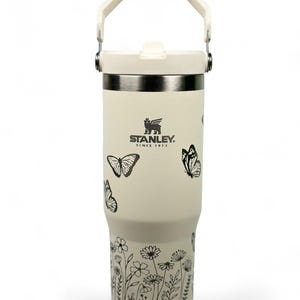 May include: Cream-colored Stanley tumbler with a white handle and lid. The tumbler features black butterfly and floral designs. The Stanley logo is printed on the front. The tumbler is designed to keep beverages cold or hot.
