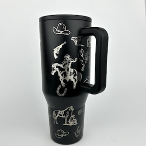 Puede incluir: Vaso t&eacute;rmico negro con asa, con dise&ntilde;os plateados grabados con tem&aacute;tica de vaqueros. Los dise&ntilde;os incluyen un vaquero a caballo, un sombrero de vaquero, un rev&oacute;lver y una herradura. El vaso tiene una tapa negra y un asa negra.