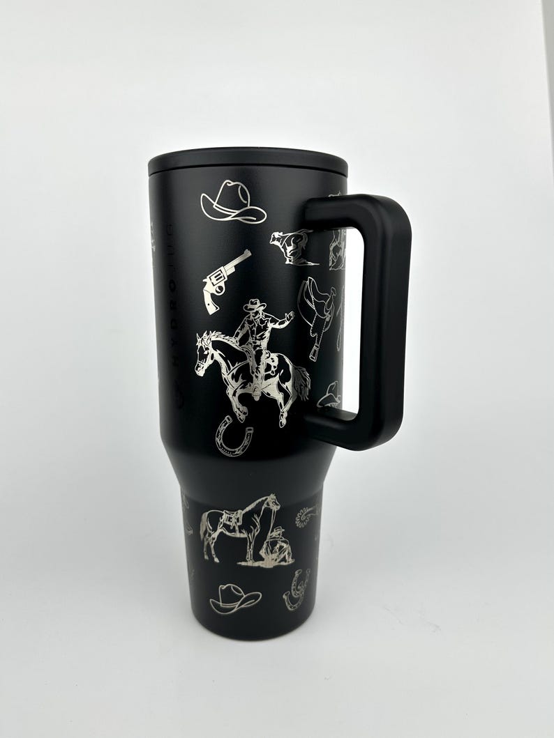 Puede incluir: Vaso t&eacute;rmico negro con asa, con dise&ntilde;os plateados grabados con tem&aacute;tica de vaqueros. Los dise&ntilde;os incluyen un vaquero a caballo, un sombrero de vaquero, un rev&oacute;lver y una herradura. El vaso tiene una tapa negra y un asa negra.