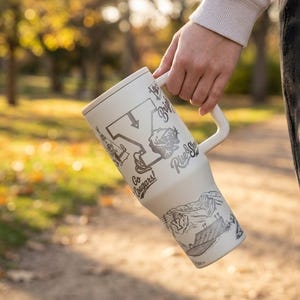 以下が含まれることがあります： クリーム色のハンドル付きトラベルマグ。黒とグレーのイラストとテキストが特徴です。マグには蓋があり、地図、カエル、「Go Cougars!」と「River Shack」の文字で装飾されています。