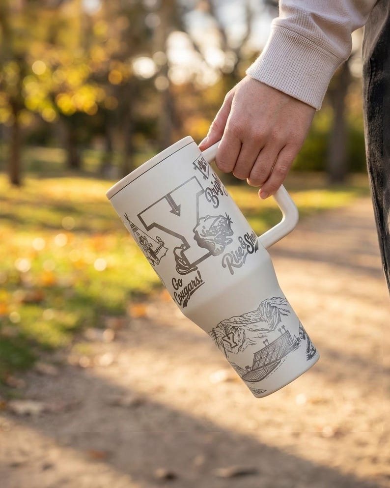 以下が含まれることがあります： クリーム色のハンドル付きトラベルマグ。黒とグレーのイラストとテキストが特徴です。マグには蓋があり、地図、カエル、「Go Cougars!」と「River Shack」の文字で装飾されています。