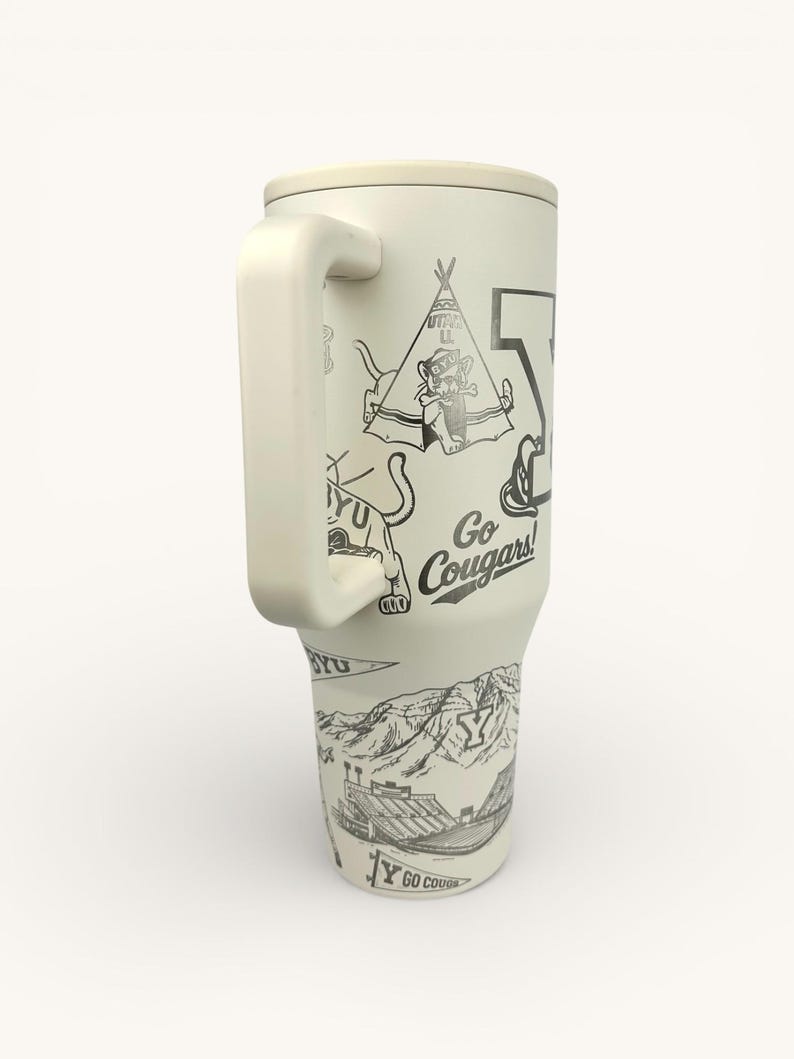 以下が含まれることがあります： 大きなハンドルが付いたクリーム色のタンブラー。ティピー、クーガー、スタジアムの黒いイラストが特徴です。「Go Cougars!」と「BYU」の文字も表示されています。実用的でスタイリッシュなドリンクウェアアイテム。