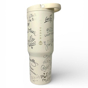 Puede incluir: Botella de agua Hydro Jug de color crema con asa y tapa de rosca. La botella est&aacute; cubierta de firmas negras de personajes de dibujos animados, como Winnie the Pooh, Tigger y Goofy. El logotipo de Hydro Jug es visible.