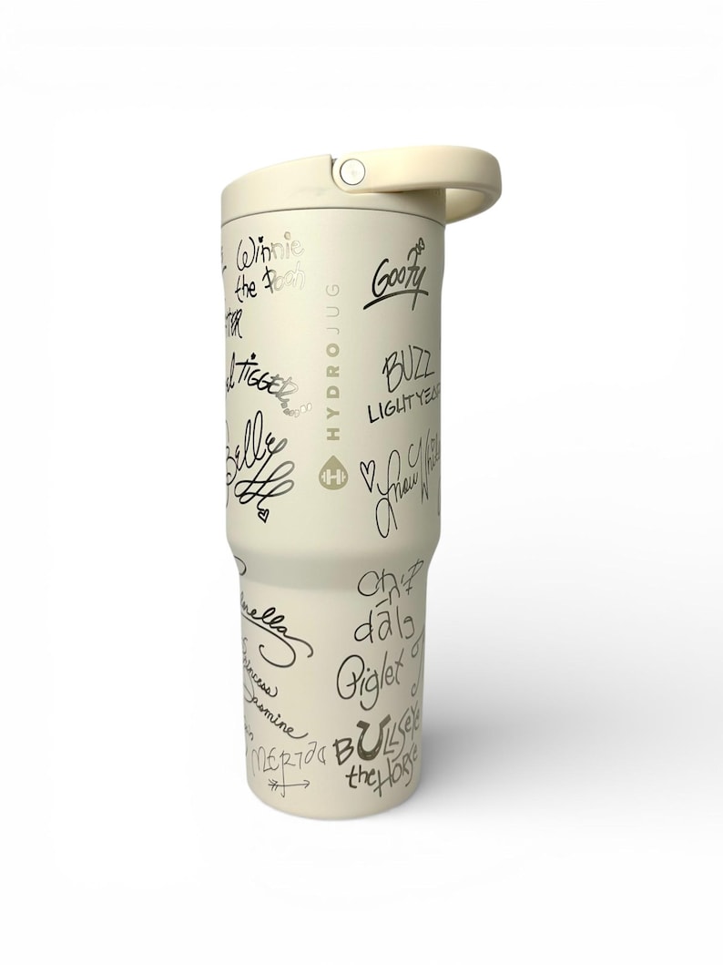 Puede incluir: Botella de agua Hydro Jug de color crema con asa y tapa de rosca. La botella est&aacute; cubierta de firmas negras de personajes de dibujos animados, como Winnie the Pooh, Tigger y Goofy. El logotipo de Hydro Jug es visible.