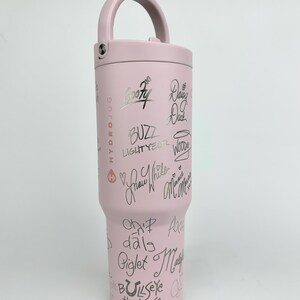 Puede incluir: Una botella de agua Hydro Jug de color rosa claro con asa y tapa de rosca. La botella est&aacute; cubierta de firmas negras y nombres de personajes, incluyendo "Goofy", "Buzz Lightyear" y "Blancanieves".