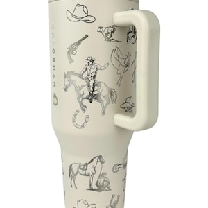 Puede incluir: Vaso t&eacute;rmico aislado de color crema con asa y tapa, con tem&aacute;tica de vaquero. El vaso est&aacute; decorado con ilustraciones en negro y plata de sombreros de vaquero, caballos, sillas de montar y un vaquero montado a caballo. El texto "HYDROJUG" est&aacute; impreso verticalmente.