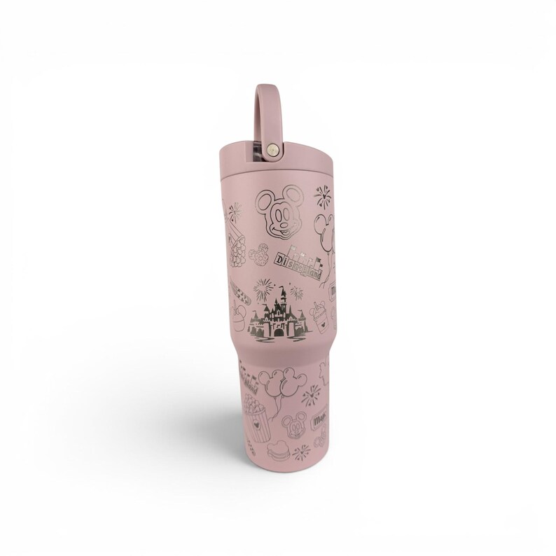 May include: A dusty rose-colored stainless steel tumbler with a matching lid and a top handle. The exterior features dark gray line art illustrations of a castle, Mickey Mouse ears, balloons, fireworks, and the text Disneyland in a repeating pattern.