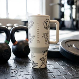 以下が含まれることがあります： ハンドルと蓋が付いた白いHydroJug。猫、フットボール選手、その他のグラフィックの白黒イラストで飾られています。ジムの環境で、質感のある黒い表面に置かれています。