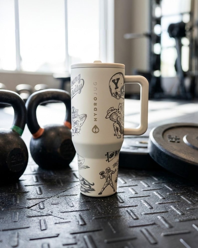 以下が含まれることがあります： ハンドルと蓋が付いた白いHydroJug。猫、フットボール選手、その他のグラフィックの白黒イラストで飾られています。ジムの環境で、質感のある黒い表面に置かれています。