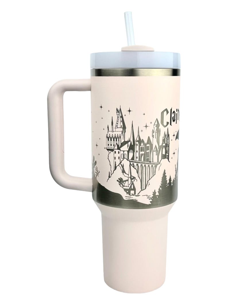 Puede incluir: Un termo de viaje rosa claro con asa, tapa blanca y pajita. El termo presenta una ilustraci&oacute;n en blanco y negro de un castillo con la palabra "Clair". Dise&ntilde;ado para mantener las bebidas fr&iacute;as o calientes.