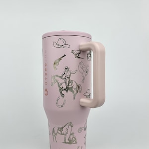 Puede incluir: Un vaso t&eacute;rmico rosa con asa y tapa, con ilustraciones plateadas de sombreros de vaquero, caballos y un vaquero. El texto "HYDROJUG" est&aacute; impreso verticalmente en el lateral. Dise&ntilde;ado para mantener las bebidas fr&iacute;as o calientes.