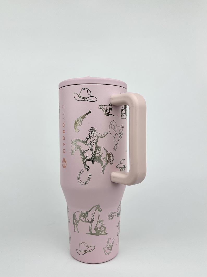 Puede incluir: Un vaso t&eacute;rmico rosa con asa y tapa, con ilustraciones plateadas de sombreros de vaquero, caballos y un vaquero. El texto "HYDROJUG" est&aacute; impreso verticalmente en el lateral. Dise&ntilde;ado para mantener las bebidas fr&iacute;as o calientes.