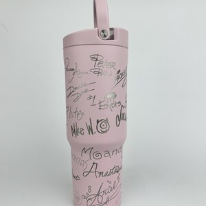 Puede incluir: Un vaso t&eacute;rmico rosa claro con asa y tapa. El vaso est&aacute; cubierto de firmas plateadas y nombres de personajes, incluyendo "Peter Pan", "Mickey" y "Ariel". El vaso est&aacute; dise&ntilde;ado para mantener las bebidas fr&iacute;as o calientes.