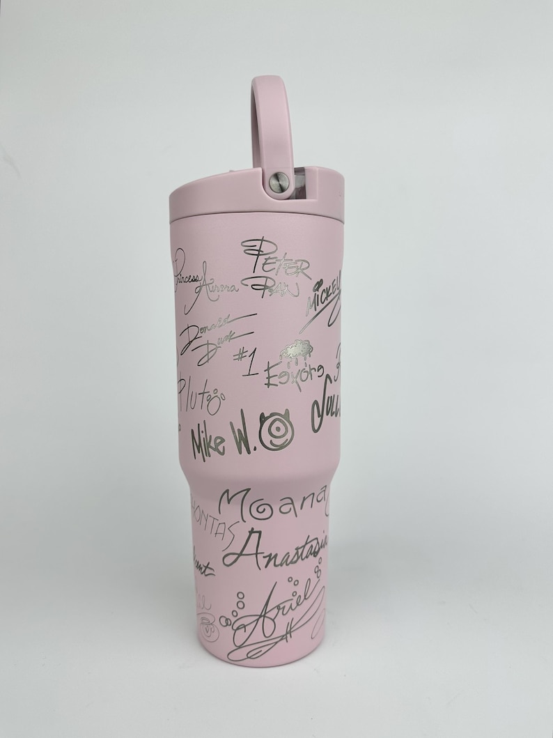 Puede incluir: Un vaso t&eacute;rmico rosa claro con asa y tapa. El vaso est&aacute; cubierto de firmas plateadas y nombres de personajes, incluyendo "Peter Pan", "Mickey" y "Ariel". El vaso est&aacute; dise&ntilde;ado para mantener las bebidas fr&iacute;as o calientes.