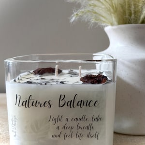 Op de afbeelding: Een heldere glazen kandelaar met een witte kaars met het opschrift "Natures Balance". De kaars heeft twee lonten en is versierd met donkerbruine stukjes. De tekst op de kaars luidt "Light a candle, take a deep breath, and feel life itself".