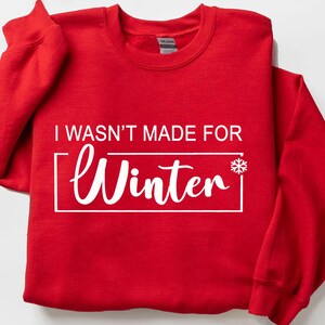 May include: A red sweatshirt with the text "I WASN'T MADE FOR Winter" in white, inside a white rectangle, with a snowflake graphic. The sweatshirt is folded and has a crew neck.