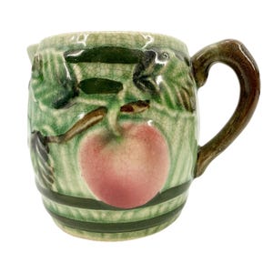 May include: A small, antique ceramic pitcher with a handle and spout. The pitcher is decorated with a pink apple and green leaves against a crackled, light green background. The handle is brown, and the base has green stripes.