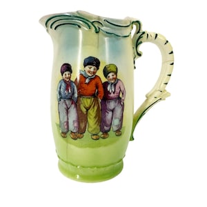 May include: A vintage ceramic pitcher with a light green base and a cream-colored body. The pitcher features a painted scene of three children in colorful clothing. The handle is cream-colored with green accents.