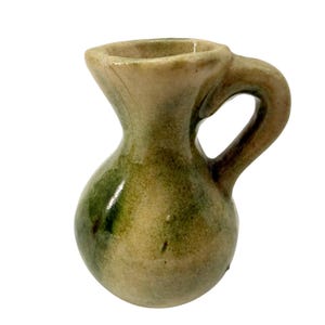 May include: A small, handcrafted ceramic pitcher with a handle. The pitcher has a bulbous body and a narrow neck, with a light beige base color and green and brown accents. The handle is curved and attached to the side of the pitcher.