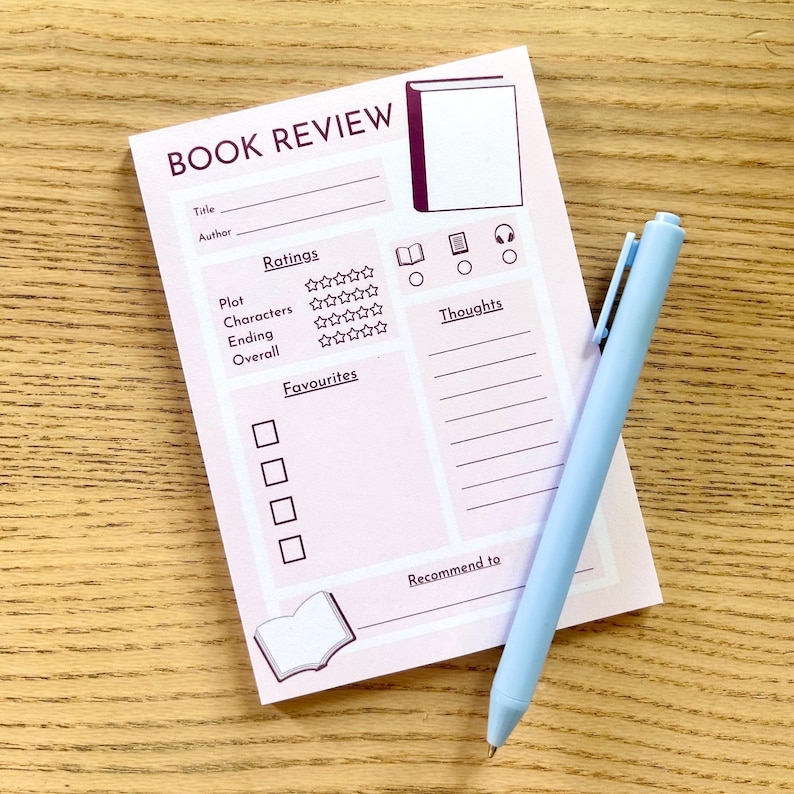 May include: A light pink book review notepad with sections for title, author, ratings, and thoughts. A light blue pen rests on the notepad. The notepad has a book graphic and the words "BOOK REVIEW".