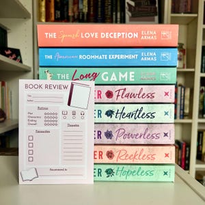 May include: A stack of colorful books with titles including "The Spanish Love Deception" and "The American Roommate Experiment." A pink book review notepad is in front of the books.