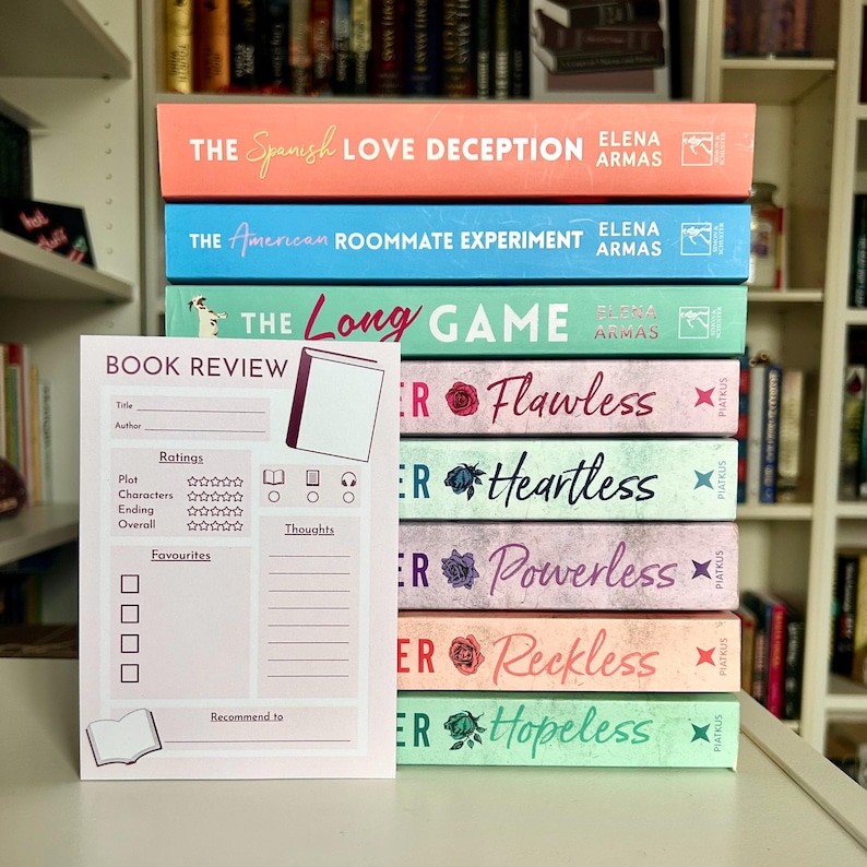 May include: A stack of colorful books with titles including "The Spanish Love Deception" and "The American Roommate Experiment." A pink book review notepad is in front of the books.
