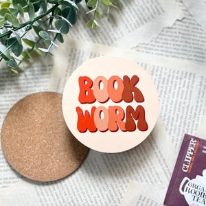 May include: A circular coaster with the words "BOOK WORM" in orange and brown, retro-style lettering. A cork coaster and a tea bag are also visible. The image is set on a background of book pages.