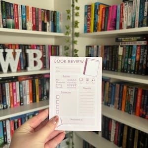 May include: A white book review card with the text "BOOK REVIEW" and rating sections. The card is held in front of a bookshelf filled with books. The card has sections for title, author, plot, characters, ending, overall, favorites, thoughts, and recommend to.
