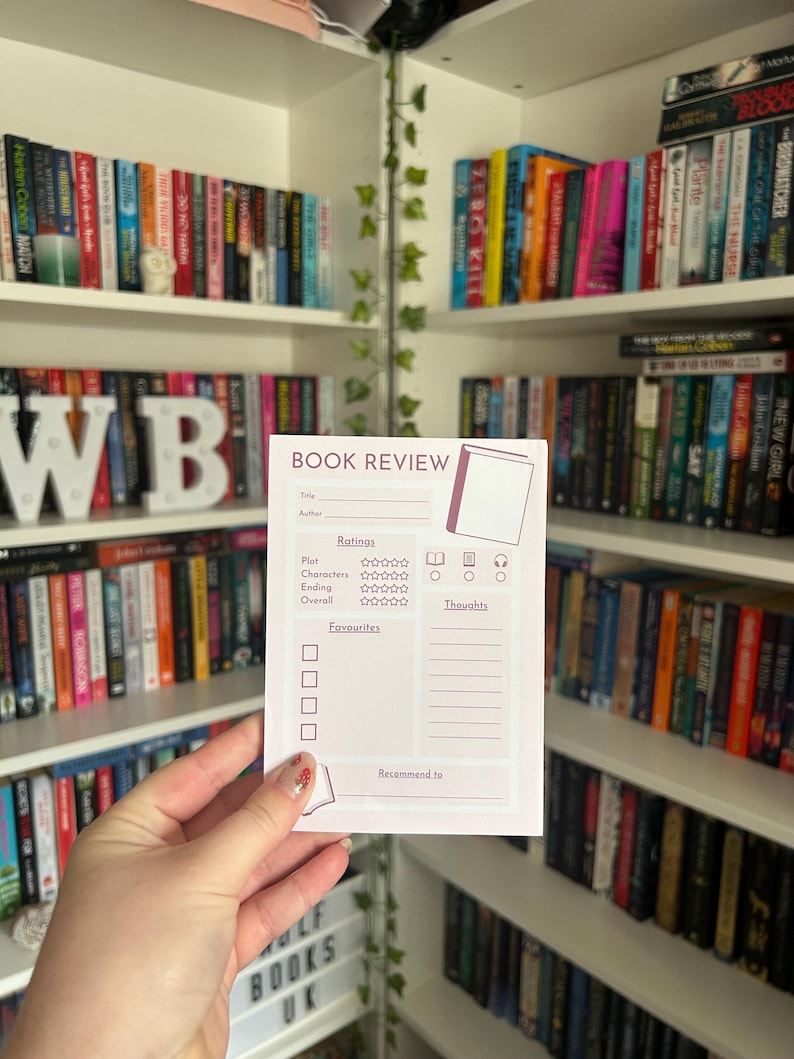 May include: A white book review card with the text "BOOK REVIEW" and rating sections. The card is held in front of a bookshelf filled with books. The card has sections for title, author, plot, characters, ending, overall, favorites, thoughts, and recommend to.