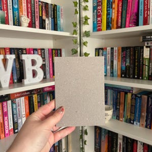 May include: A square, light gray piece of cardboard is held in front of a bookshelf filled with colorful books. The books are arranged on white shelves, and some have decorative covers. The background includes a decorative vine.
