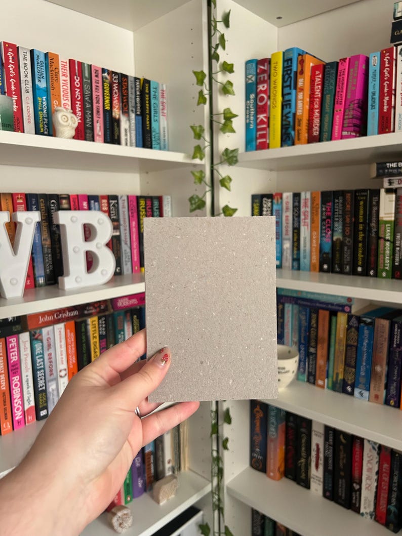 May include: A square, light gray piece of cardboard is held in front of a bookshelf filled with colorful books. The books are arranged on white shelves, and some have decorative covers. The background includes a decorative vine.