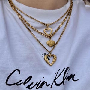 May include: Three gold-colored necklaces with heart pendants. The necklaces are layered, with different chain styles. The person is wearing a white t-shirt with the text "Calvin Klein New York" in black script.