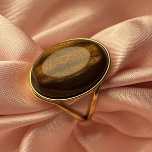 May include: A gold ring with a large oval-shaped tiger's eye gemstone. The ring is set on a soft pink fabric background.