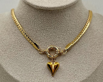 May include: A gold-colored necklace with a heart-shaped pendant. The necklace features a herringbone chain and a circular clasp. The pendant is a solid, pointed heart. The necklace is displayed on a mannequin head against a white background.
