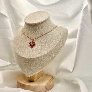 May include: A gold-colored necklace with a red heart-shaped pendant featuring an eye design. The pendant is displayed on a mannequin. The background is a white, draped fabric.