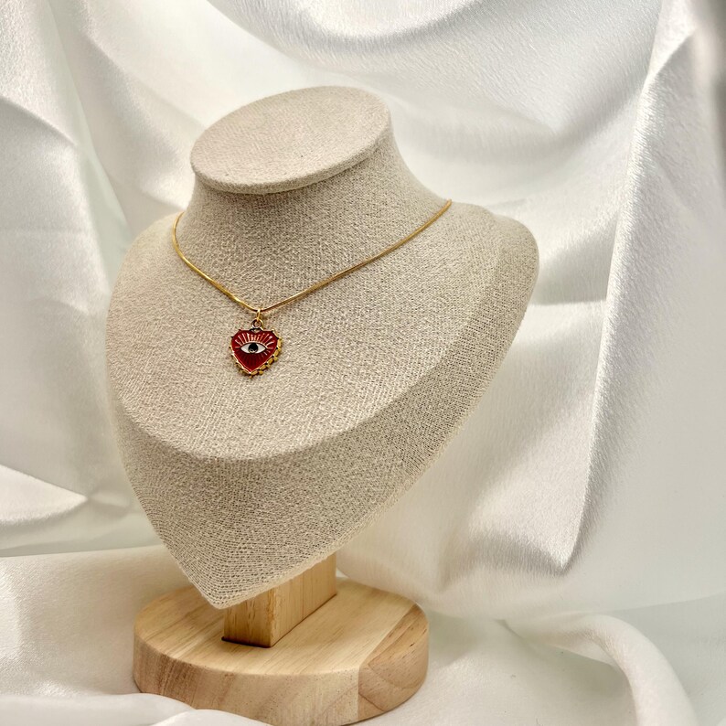 May include: A gold-colored necklace with a red heart-shaped pendant featuring an eye design. The pendant is displayed on a mannequin. The background is a white, draped fabric.